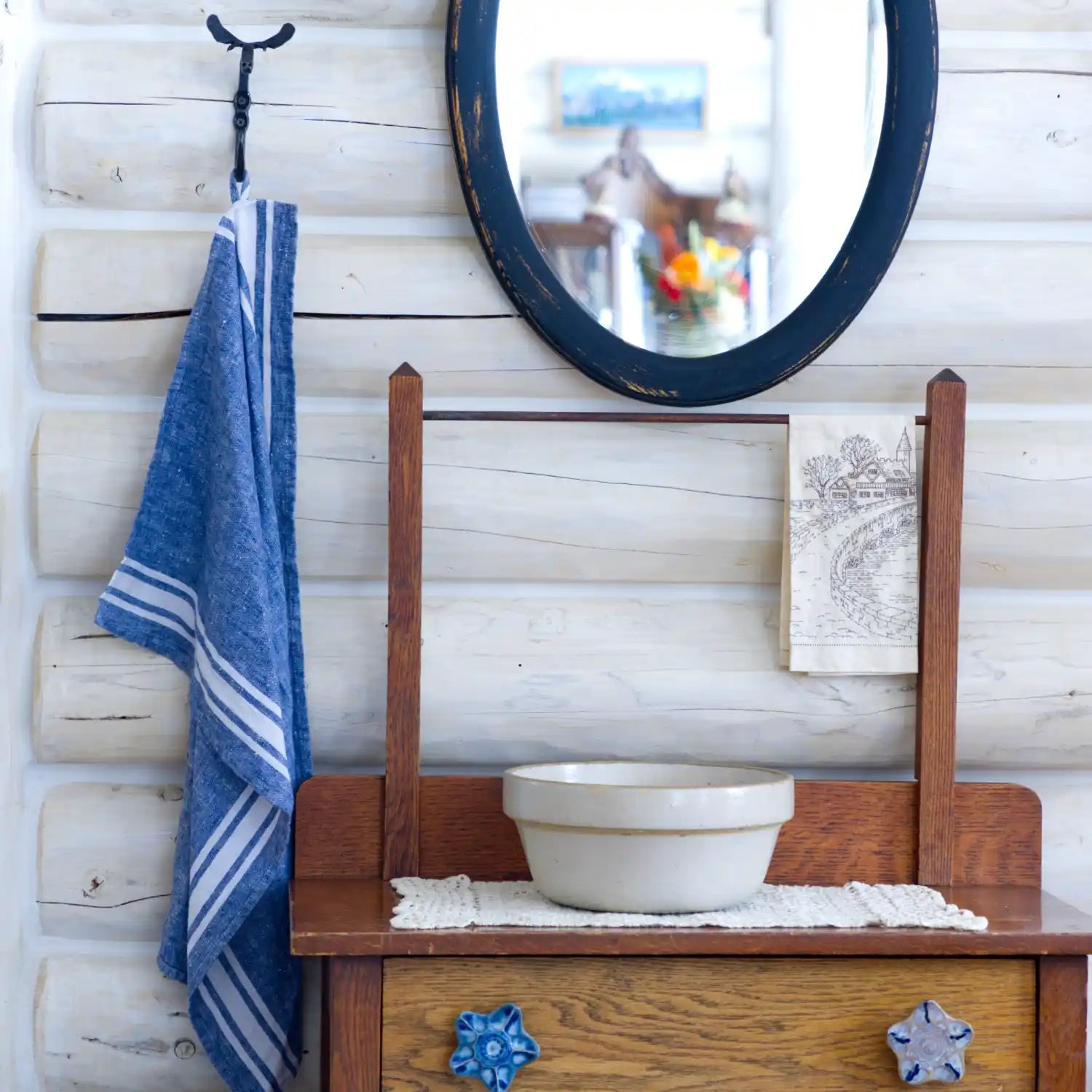 Blue and white striped flax linen hand towel hanging from blacksmith hook in log house with a mirror and washstand beside.